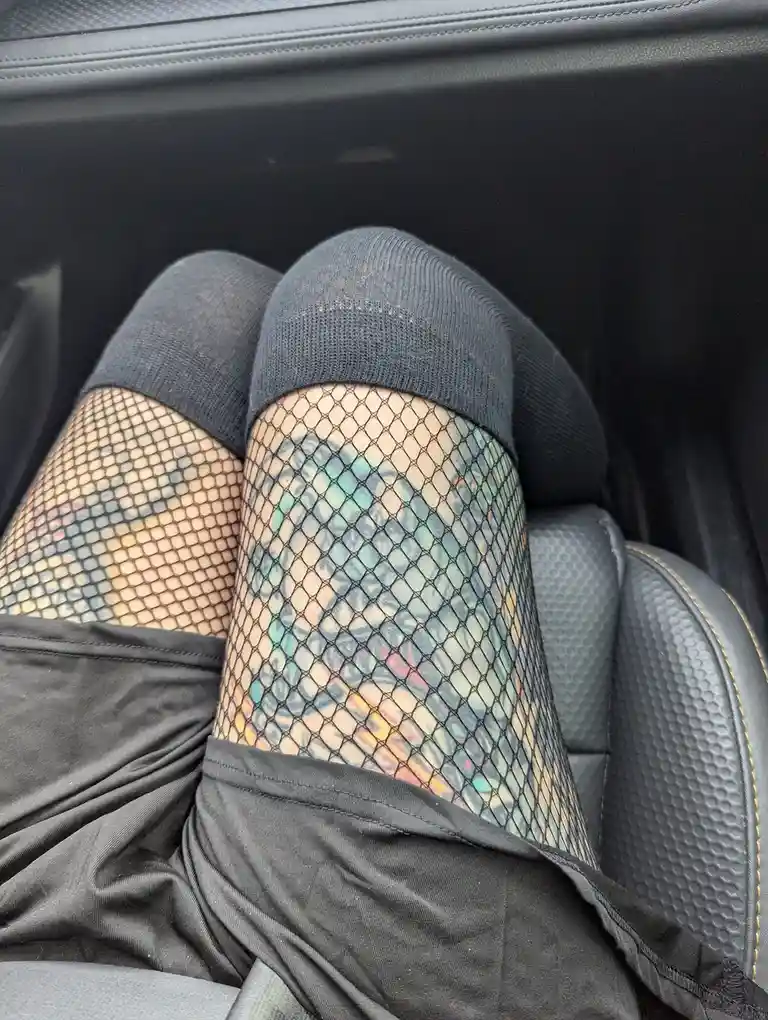 A close-up photo shows a person's tattooed legs inside a car, wearing black fishnet stockings over dark socks. The tattoos…