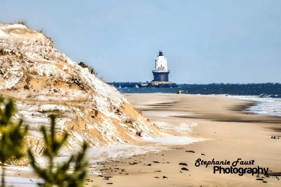 A scenic beach photo shows a sandy shore with a large dune on the left covered in patches of snow, leading to a distant…