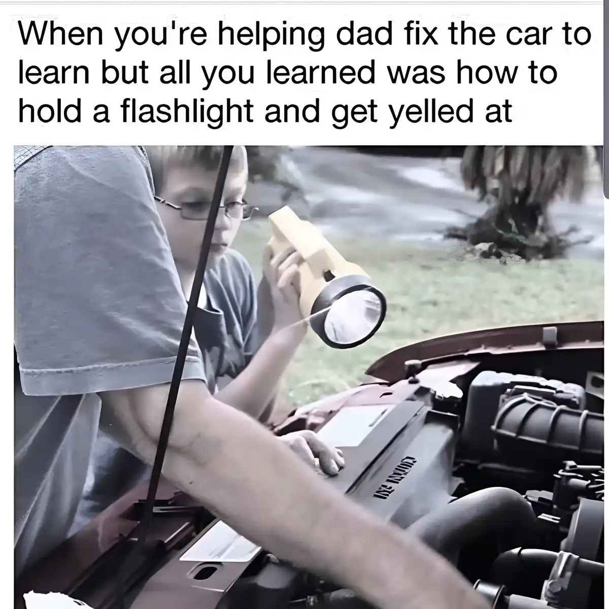 A young boy with glasses holds a flashlight, shining it into the engine bay of a car with its hood open, while an adult's…