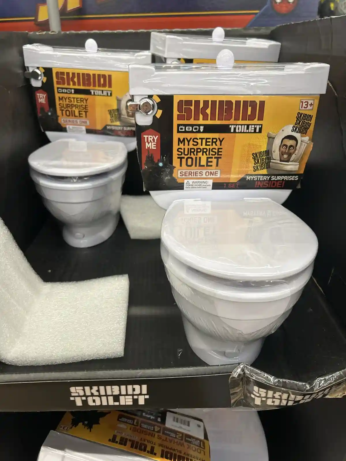 Two white toy toilets in retail packaging are displayed on a shelf, labeled 'SKIBIDI TOILET MYSTERY SURPRISE TOILET SERIES…