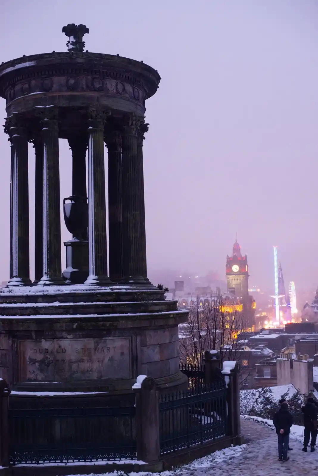A snowy, atmospheric photo of the Dugald Stewart Monument in Edinburgh, with a cityscape in the background featuring a clock…