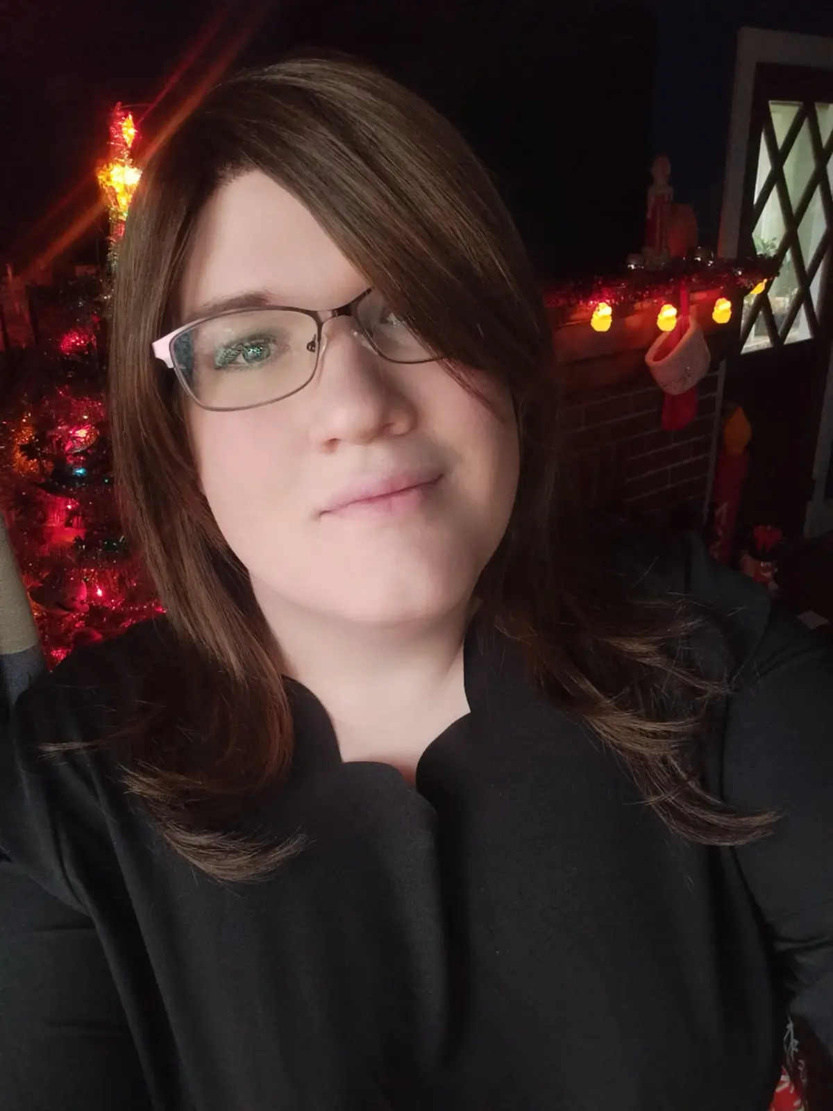 A person with brown hair and glasses, wearing a black shirt, looks at the camera with a slight smirk while standing in front…