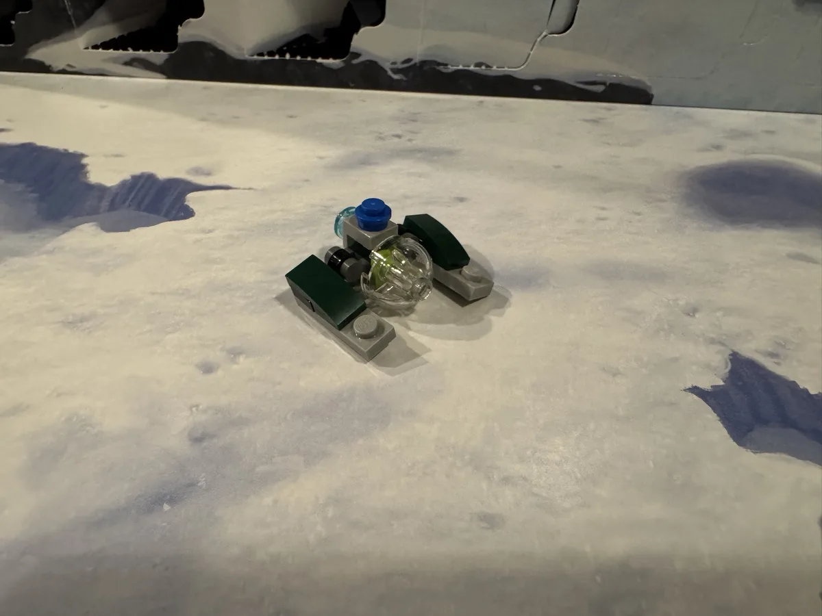 A small, crudely assembled Lego vehicle with green, gray, and transparent pieces sits on a surface painted to resemble the…