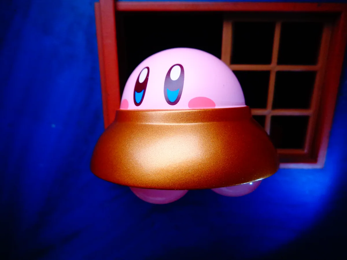 A pink, round, cartoon character with large blue eyes and rosy cheeks peeks out from a golden UFO-shaped object, set against…