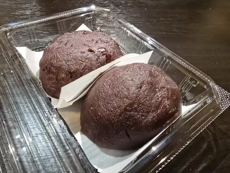Two dark brown, round, baked goods resembling chocolate muffins sit side by side in a clear plastic takeout container lined…