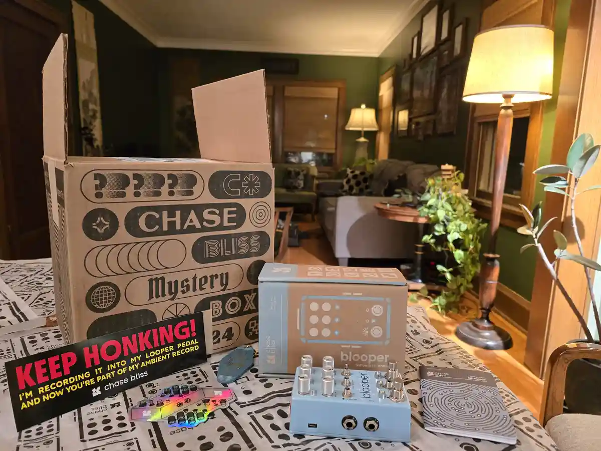 A Chase Bliss Mystery Box sits open on a table with its contents, including a blue 'blooper' looper pedal, a manual, and a…