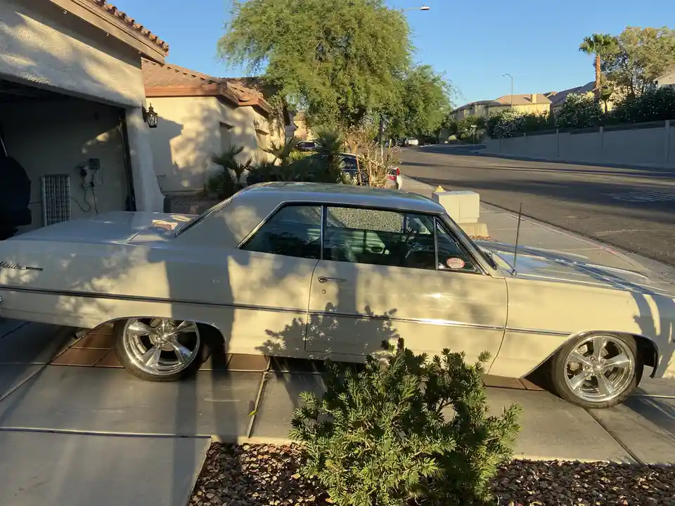 A cream-colored classic Chevrolet Malibu is parked in a driveway, its shiny chrome wheels and body reflecting sunlight. The…