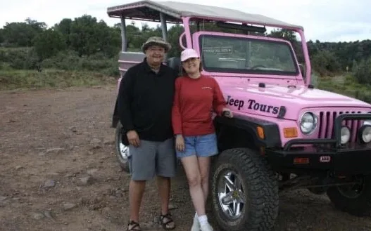 A man and a woman stand smiling in front of a pink Jeep Wrangler with 'Jeep Tours' written on the side. The vehicle has a…