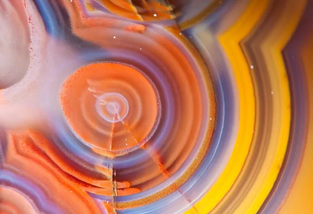 A close-up photo of a colorful, swirling agate stone with concentric orange, yellow, and purple bands. Overlaid text reads…