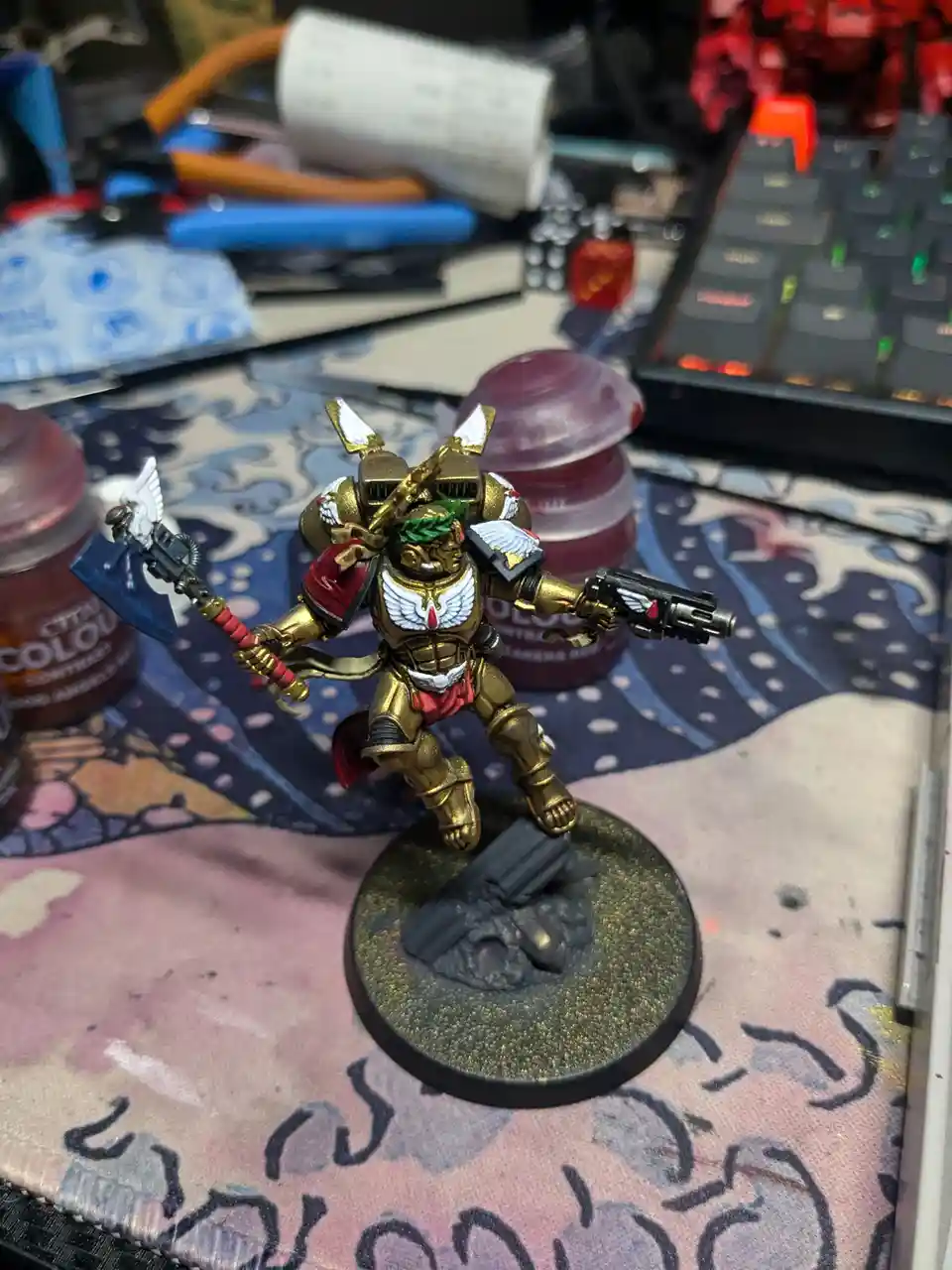 A painted miniature of a Space Marine from Warhammer 40,000, wearing gold armor with white wings and red accents, stands on…