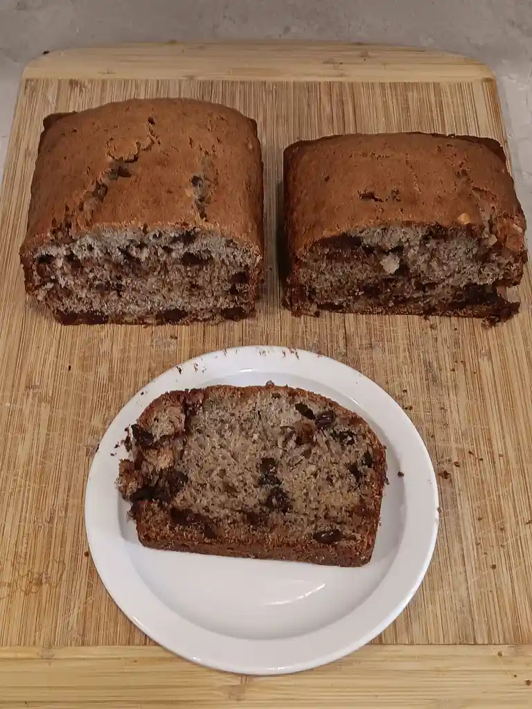 Two loaves of banana bread, sliced, sit on a wooden cutting board, with one slice served on a white plate in the foreground…