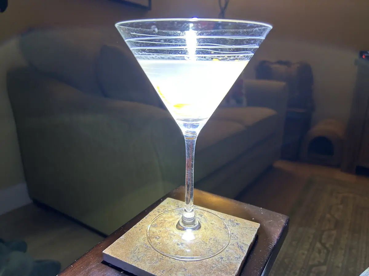 A close-up photo shows a martini glass filled with a clear liquid and a lemon twist, sitting on a coaster atop a wooden…
