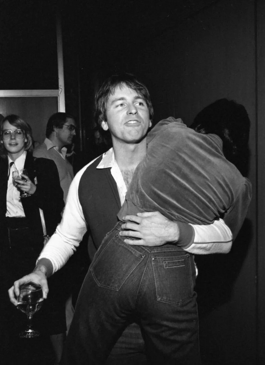 A black-and-white photo shows a man in a white shirt and dark vest holding another person in a tight embrace from behind…