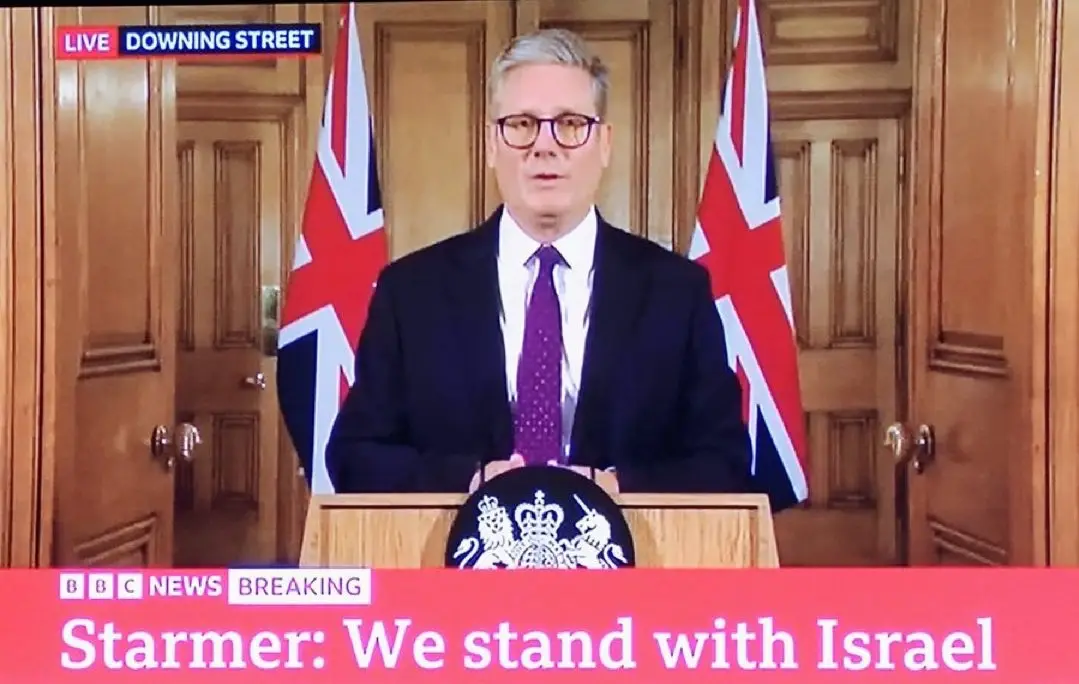 A man in a suit and glasses stands at a podium with the British coat of arms, flanked by two Union Jack flags, in what…