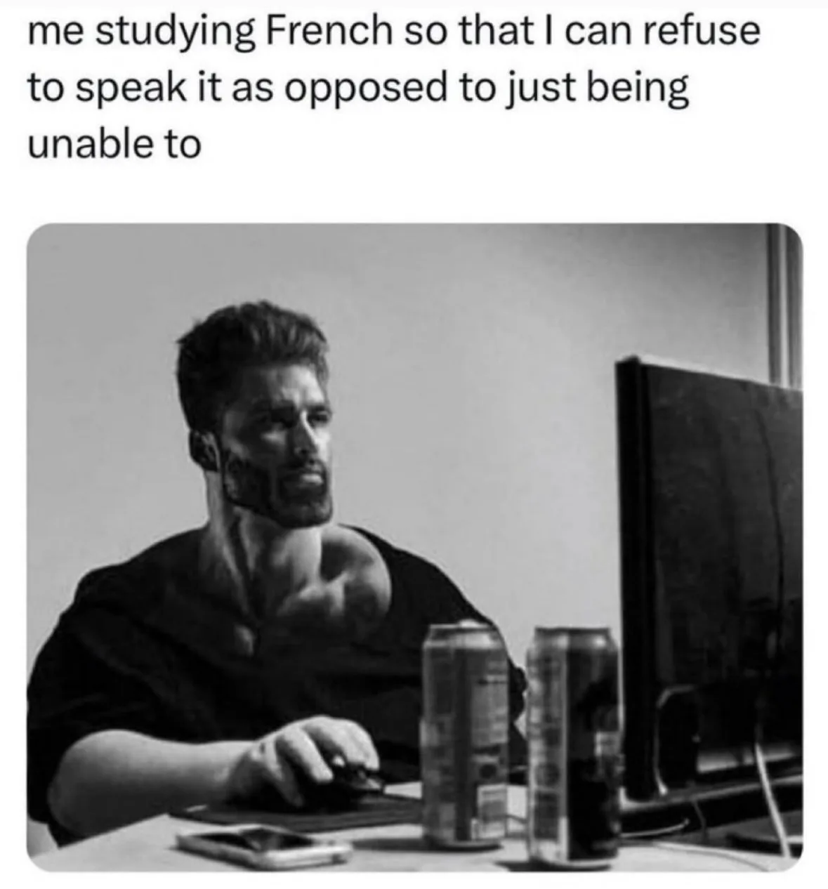 A black-and-white photo shows a man with a beard sitting at a desk, using a computer mouse. Two soda cans and a phone are on…