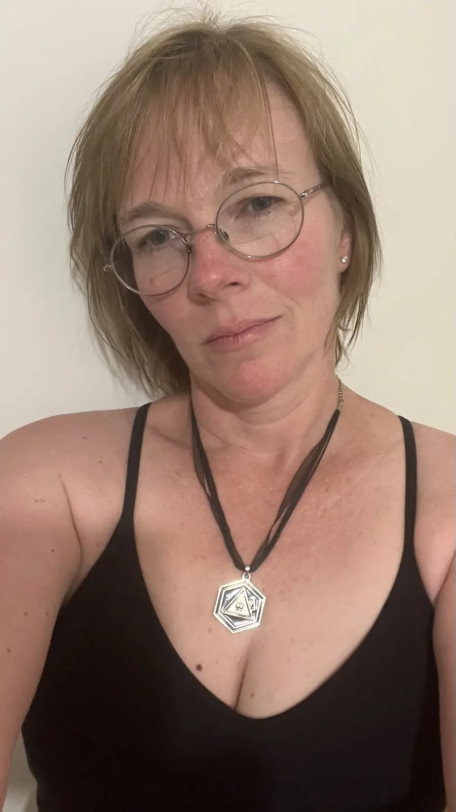 A woman with short blonde hair and round glasses wears a black tank top and a necklace with a hexagonal pendant featuring a…