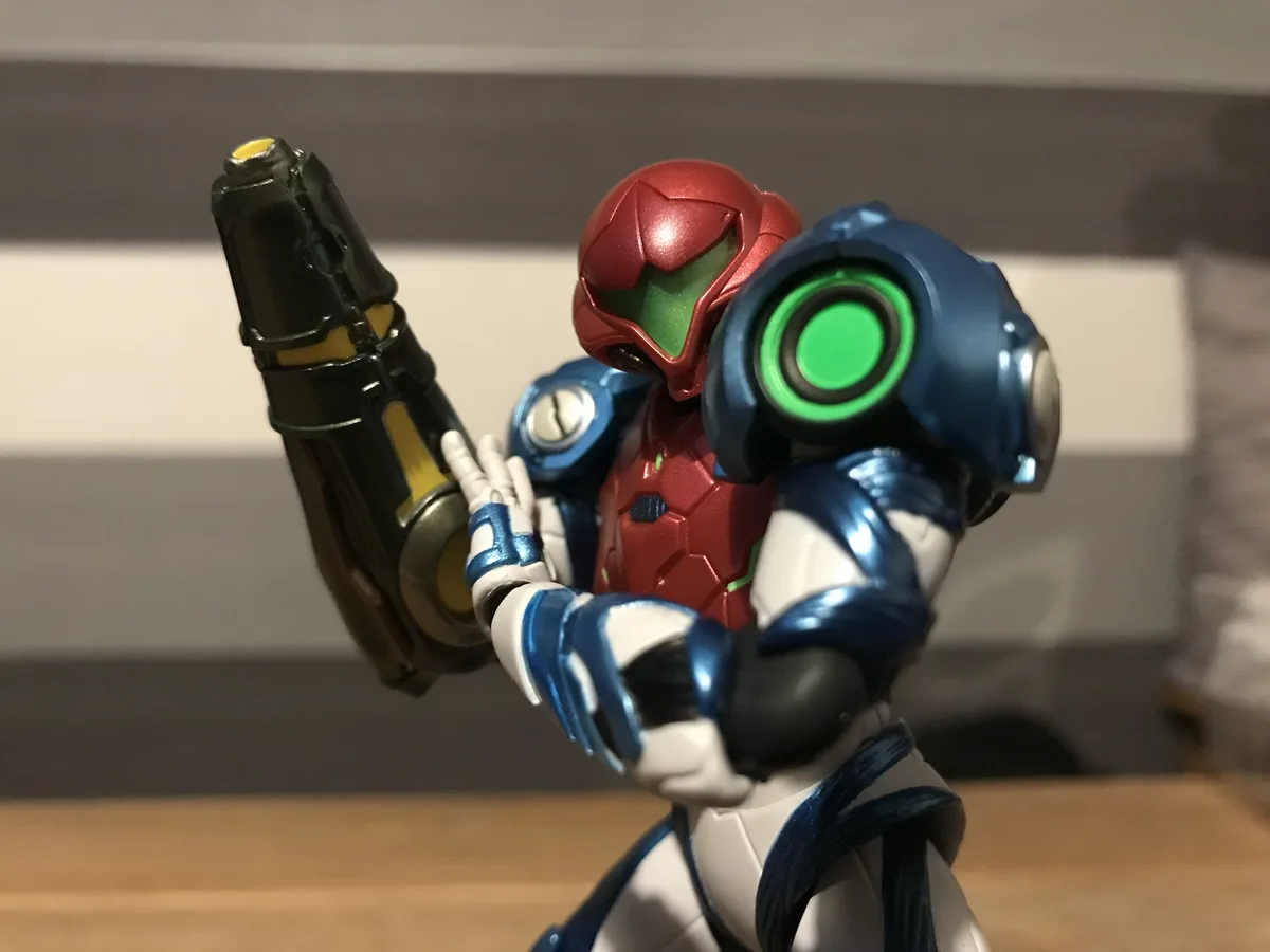 A close-up photo of a Samus Aran action figure from the Metroid series, posed holding a weapon with her left hand while her…
