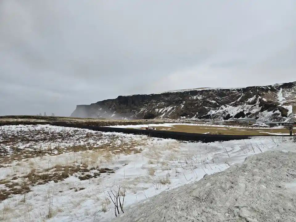 A snowy, desolate landscape with a black road cutting through snow-covered fields, leading toward distant cliffs and sparse…