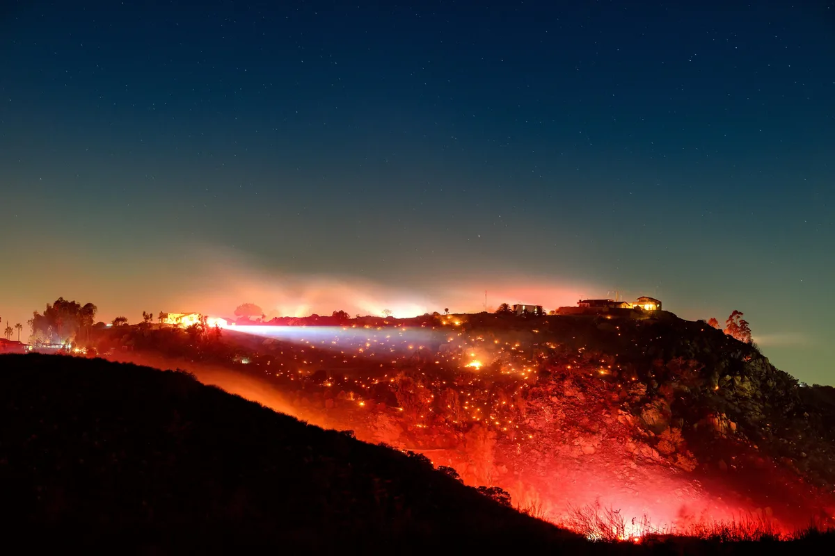 A nighttime photograph shows a hillside engulfed in wildfire, with glowing embers and flames illuminating the terrain under…