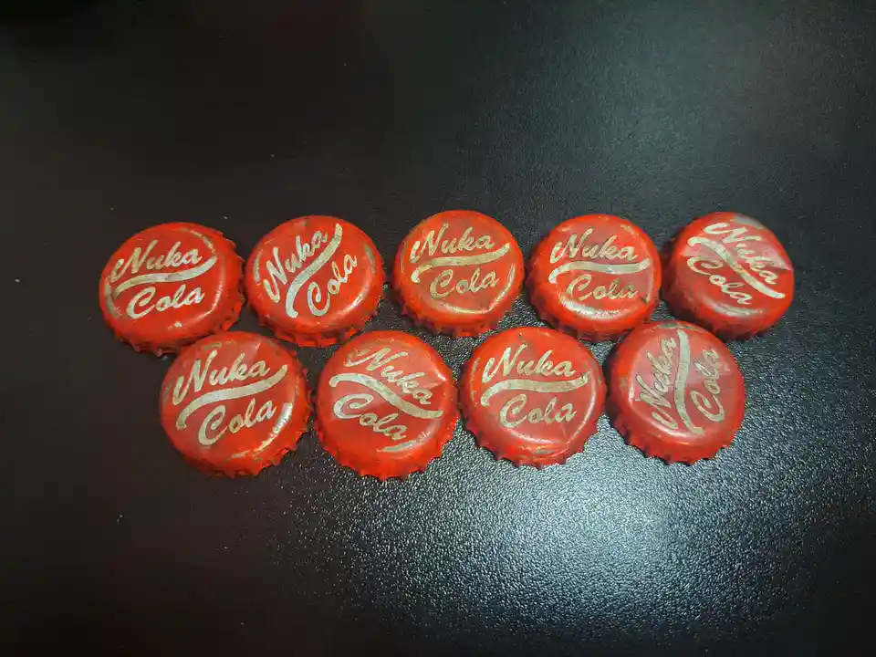 Nine red, circular Nuka-Cola bottle caps with gold lettering are arranged in two rows on a dark, textured surface. The caps…