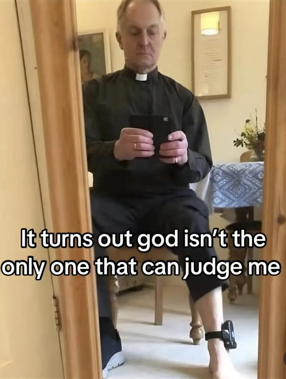 A man dressed in a black clerical collar shirt sits on a wooden chair, looking down at his phone. He has a small black…