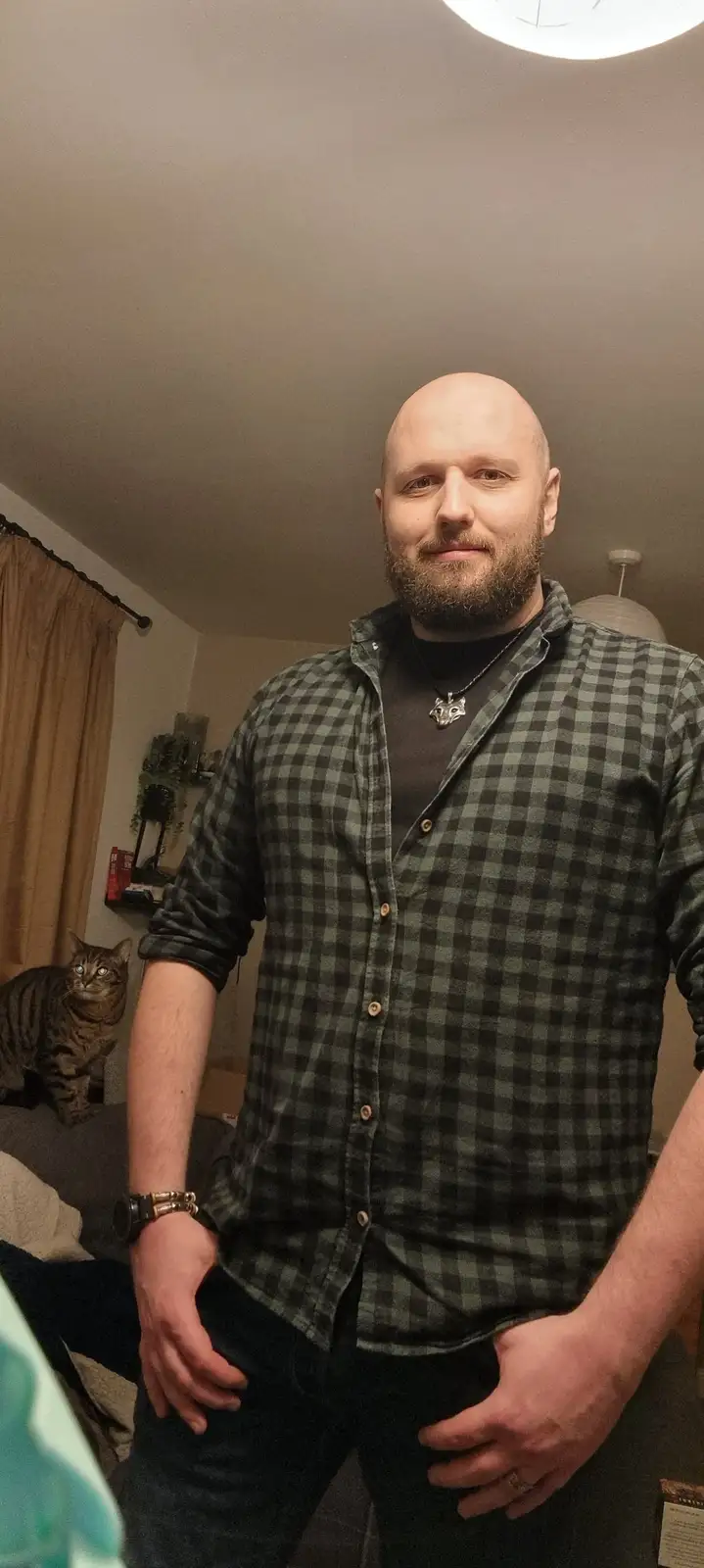 A bald man with a beard wearing a green plaid shirt and a wolf pendant necklace stands in a room with his hands in his…
