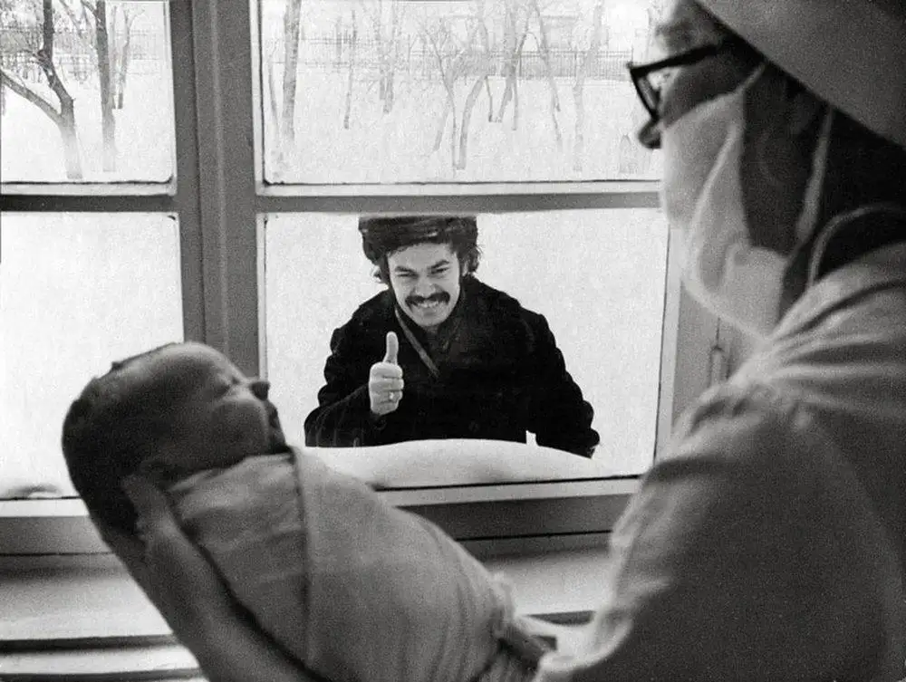 A black-and-white photo shows a man with a mustache giving a thumbs-up through a window while a newborn baby is held by a…