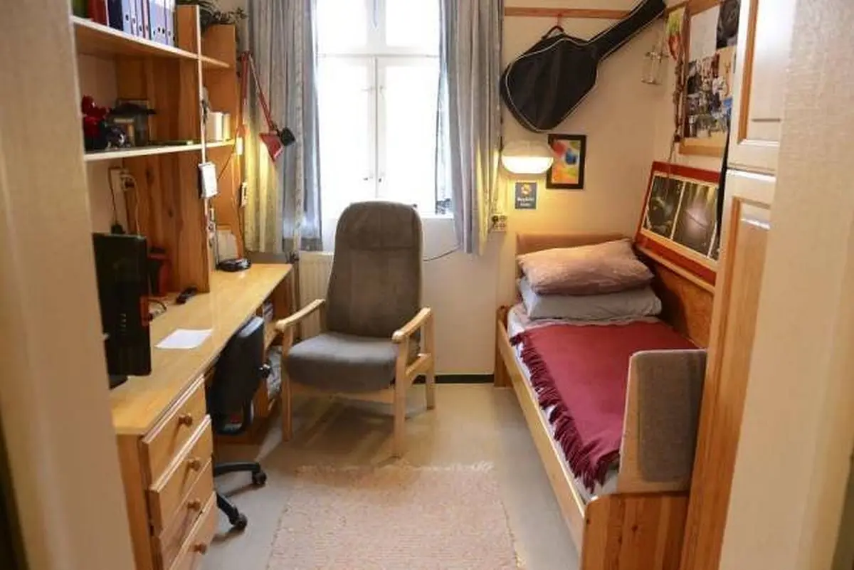 A narrow, cozy dorm room with a wooden desk, chair, and bed against the wall. A guitar case hangs on the wall above the bed…