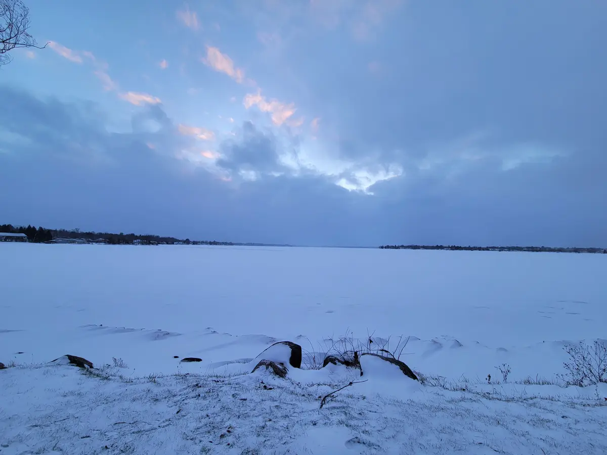 A wide, serene winter landscape shows a frozen lake covered in snow under a twilight sky with soft pink and blue clouds. In…