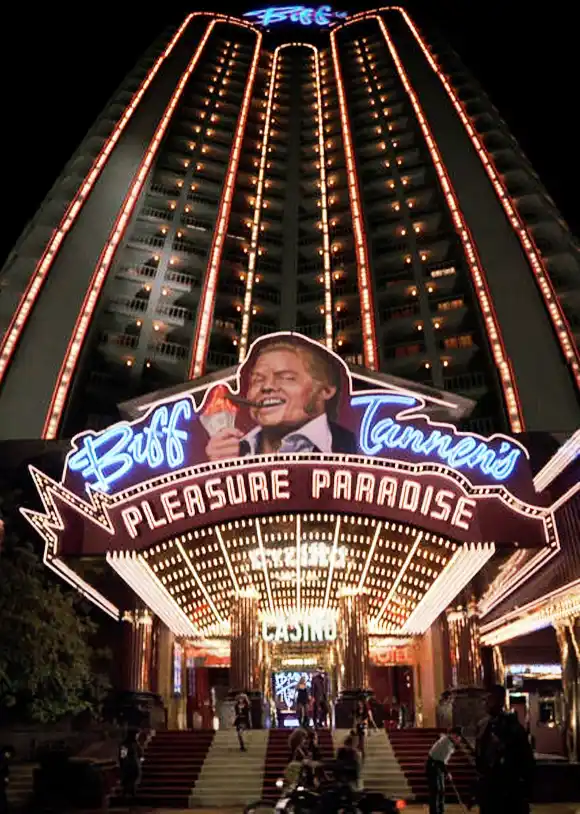 A nighttime photo of a tall, brightly lit casino building with a large, glowing marquee. The marquee features a smiling man…