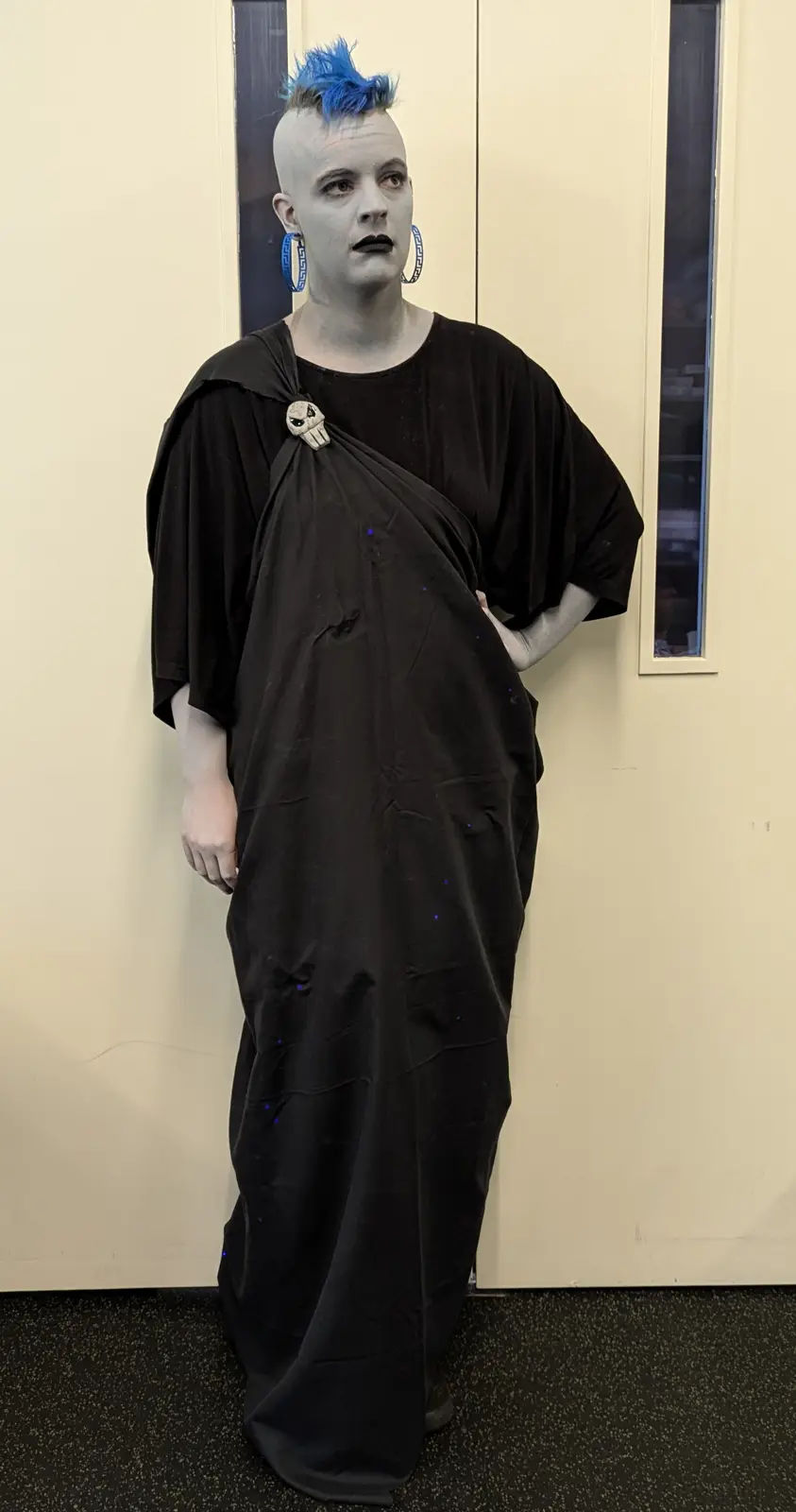 A person with a shaved head, blue mohawk, white face paint, and large hoop earrings stands in a black draped robe resembling…