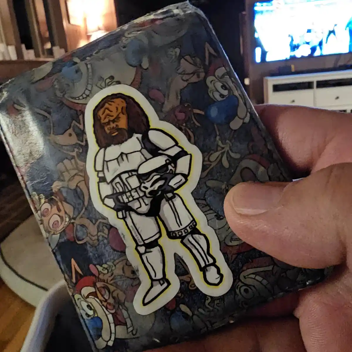 A hand holds a wallet covered in cartoon stickers, prominently featuring a sticker of a stormtrooper with a long beard and…