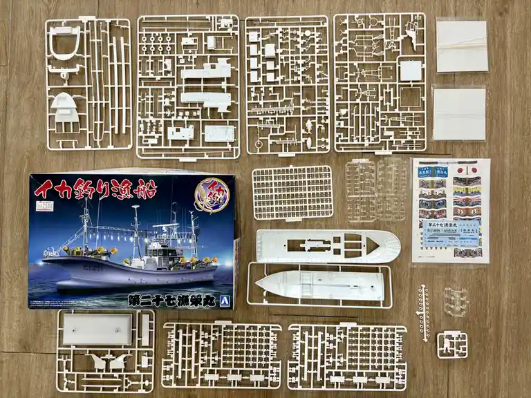An overhead shot displays the contents of a plastic model kit for a Japanese squid fishing boat, laid out on a wooden floor…
