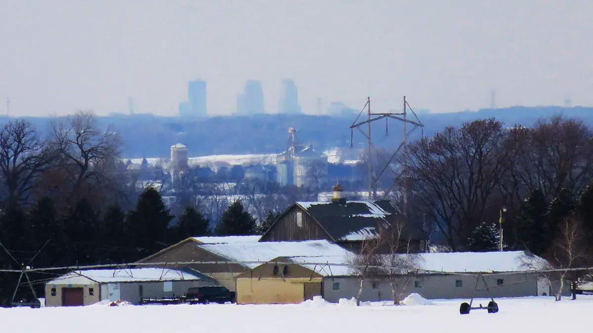 A snowy rural landscape with farm buildings in the foreground and a hazy city skyline visible in the distance. The overlaid…