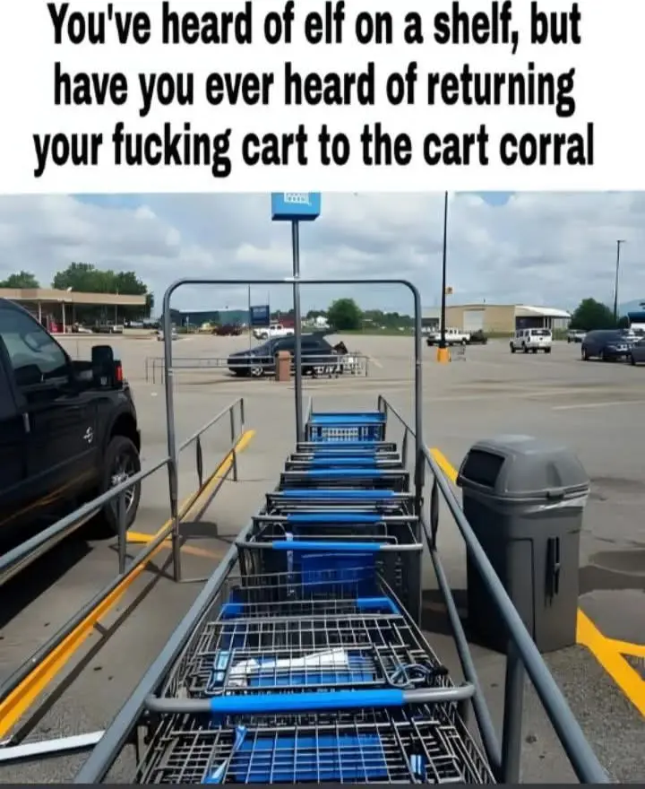 A row of blue shopping carts is lined up in a metal cart corral at a store's parking lot, with a trash can and a black truck…