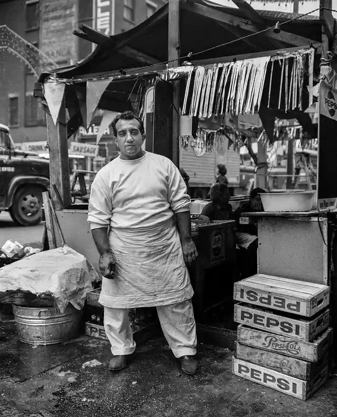 A black-and-white photo shows a man in a stained apron standing in front of a street food cart with stacked Pepsi crates…