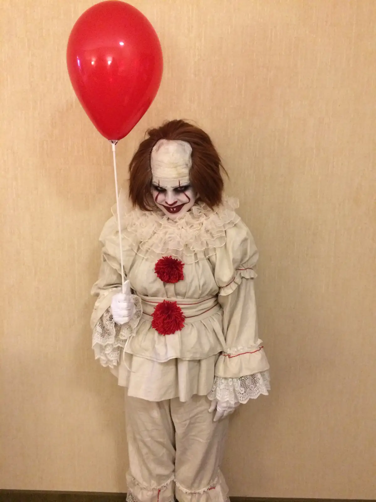 A person in a Pennywise the Clown costume from 'It' holds a red balloon, wearing a white ruffled outfit with red pom-poms…