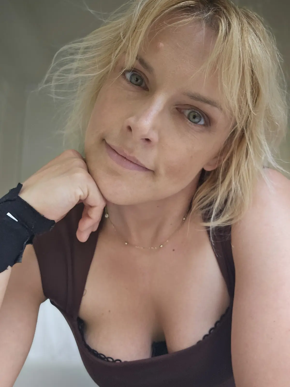 A woman with blonde hair and green eyes looks directly at the camera with a slight smile while resting her chin on her hand…