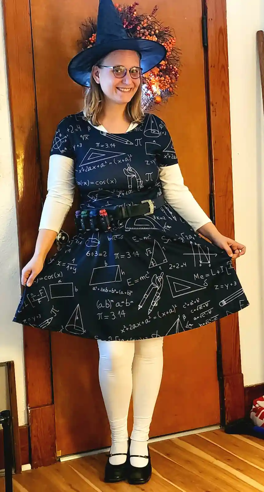 A smiling woman in a witch costume, wearing a blue pointed hat and a black dress covered in white mathematical equations and…