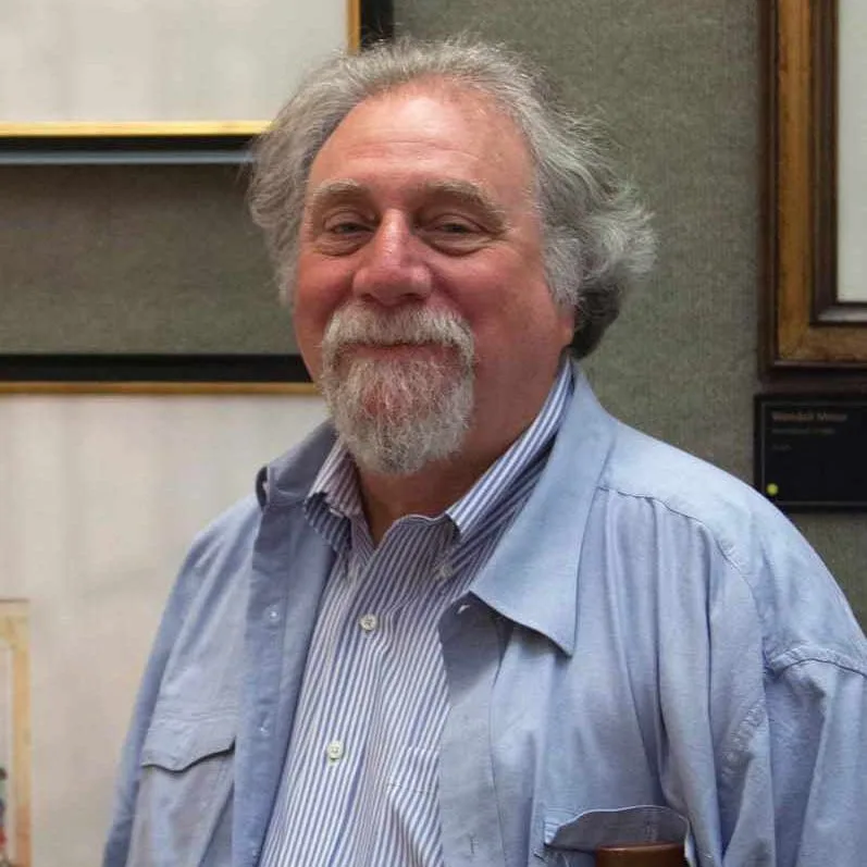 a smiling older man with gray hair and a beard, wearing a light blue button-up shirt over a striped collared shirt, stands…