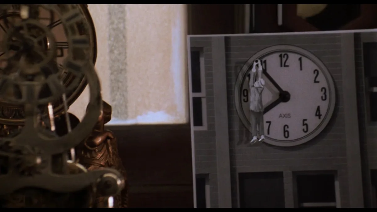 A still from a movie shows a large, ornate clock on the left and a cardboard cutout of a building with a clock face on the…