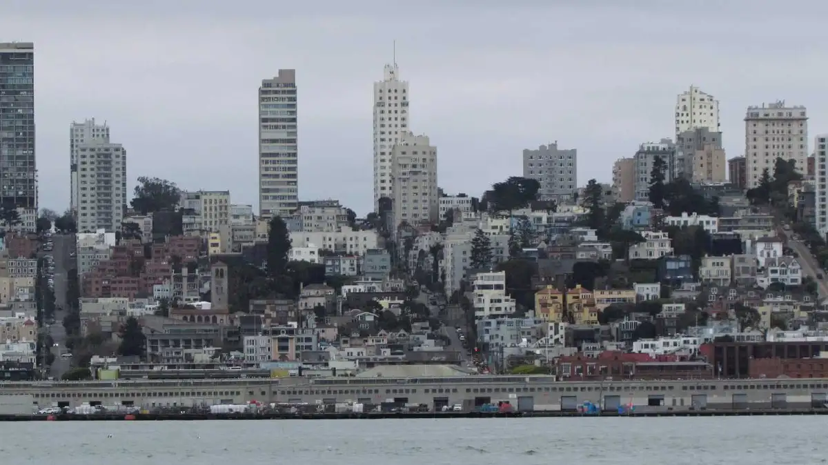 A wide shot of a hilly cityscape with numerous buildings of varying heights and colors, viewed from across a body of water…