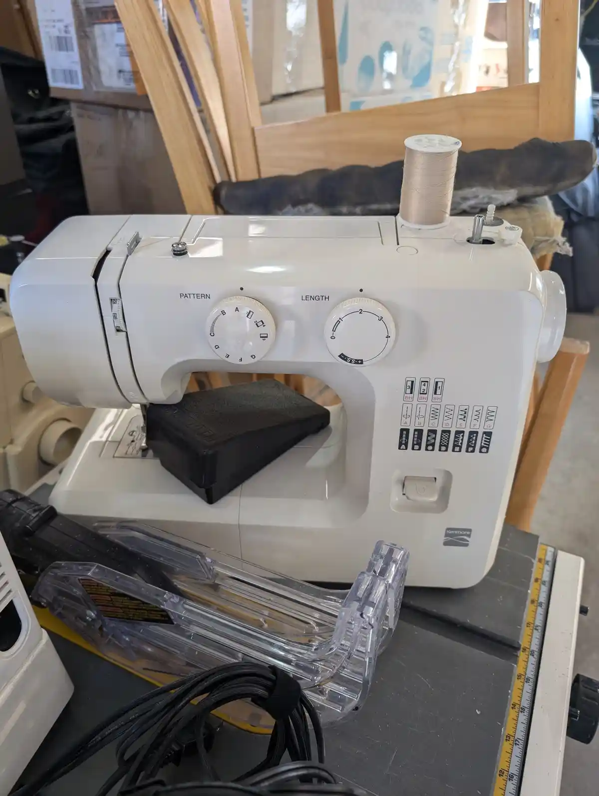 A white Kenmore sewing machine sits on a workbench, with a spool of beige thread on top and various tools and cables nearby…