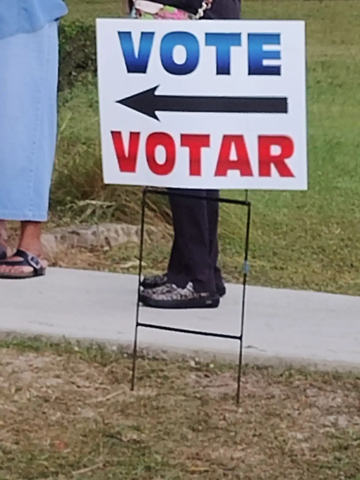 A white yard sign with a black arrow pointing left displays 'VOTE' in blue text at the top and 'VOTAR' in red text at the…