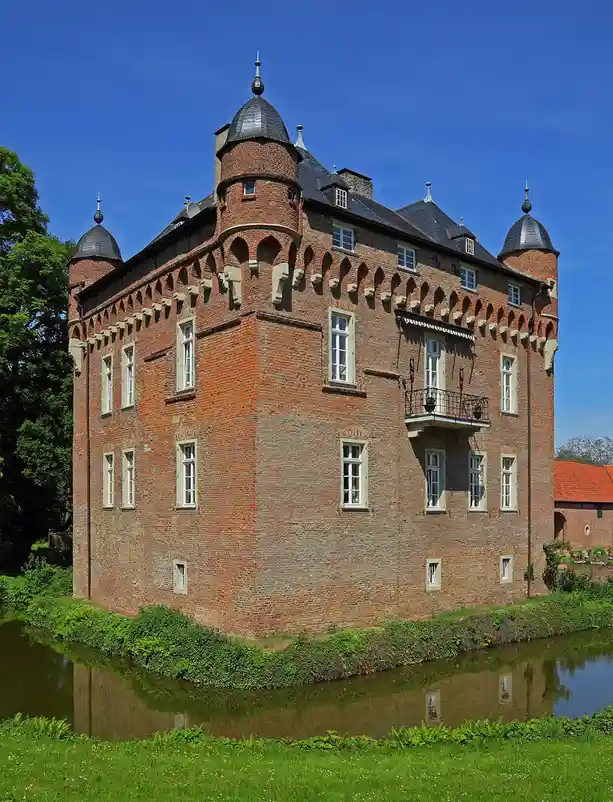 A grand, red-brick castle with multiple turrets and a dark roof stands beside a moat under a clear blue sky. The overlaid…