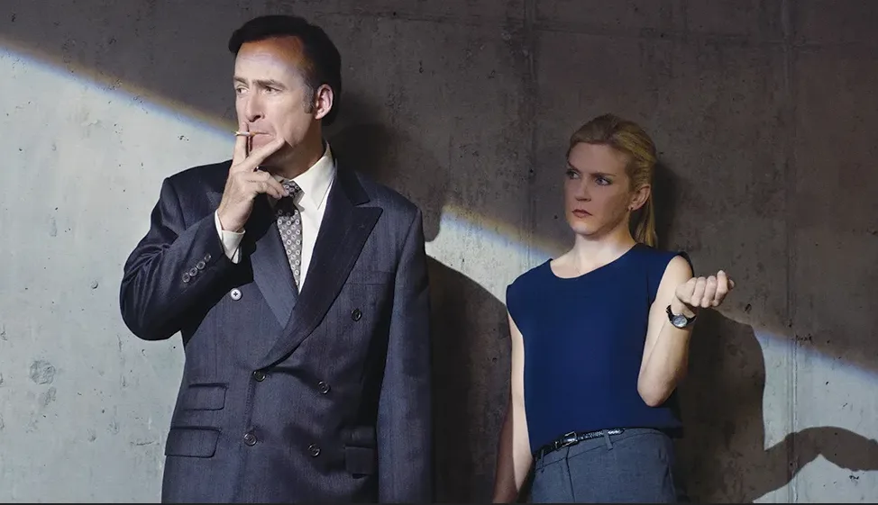 a man in a dark suit holding a cigarette to his lips while looking off to the side, standing next to a woman in a blue top…