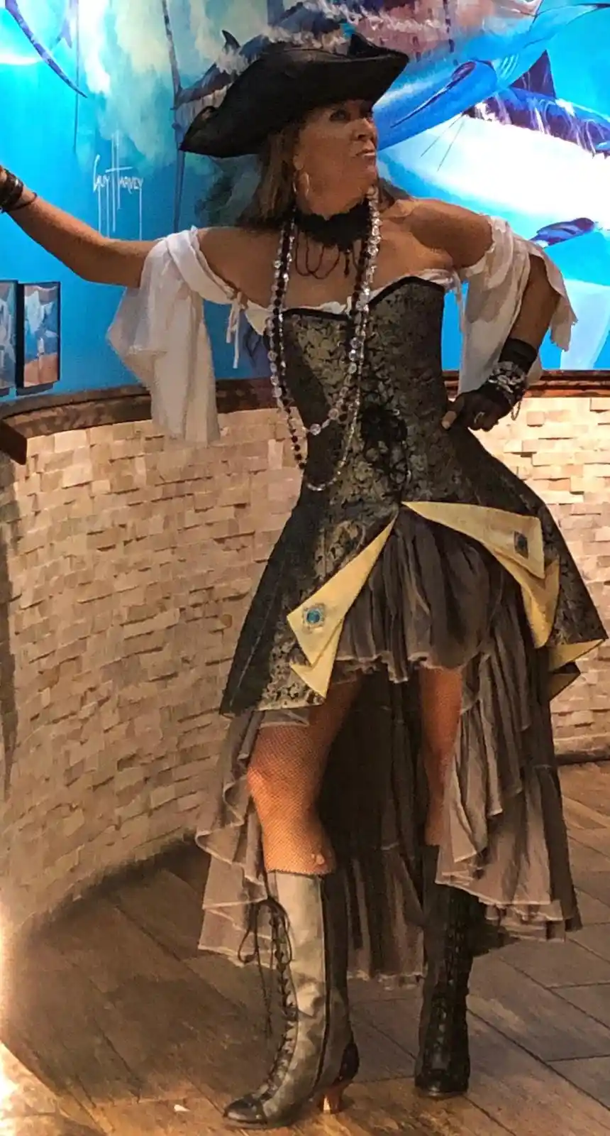 A woman in a pirate costume with a black tricorn hat, corset, layered skirt, and boots poses with one arm raised. The…