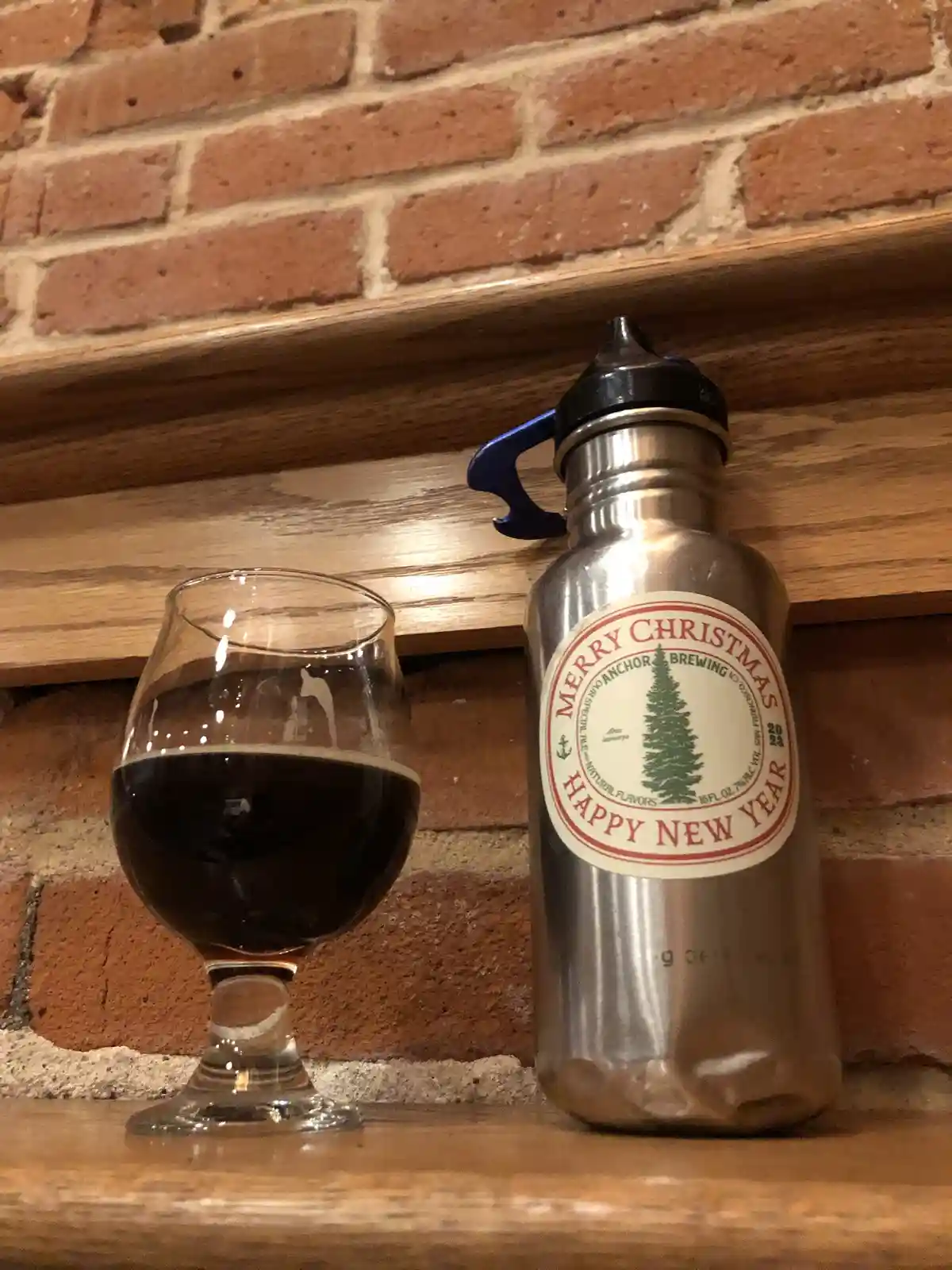 A stainless steel water bottle with a 'Merry Christmas Happy New Year' label from Anchor Brewing sits next to a glass of…