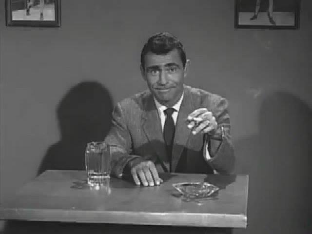 A black-and-white image of a man in a suit sitting at a table with a glass of water and an ashtray, smiling smugly while…
