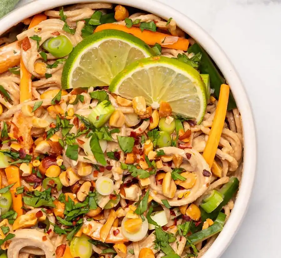a close-up photo of a white bowl filled with peanut noodles, garnished with lime wedges, chopped peanuts, sliced green…
